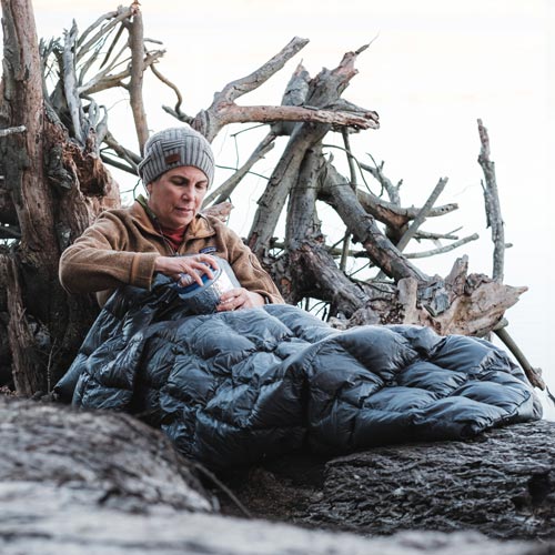 Woman backpacker enjoying coffee on log snuggled in a Stratum 55 Degree Top Quilt